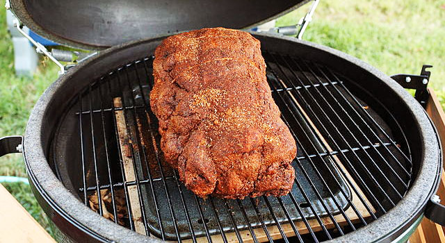 Pork shoulder rubbed with spices on a grill grate with a deflector and a drip pan between the meat and the coals