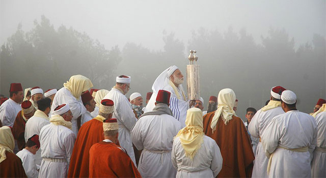 A congregation in robes and headwear gathers around an older man dressed in white holding a large scroll vertically in front of him, with trees and mist in the background