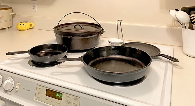 A cast iron Dutch oven, small fry pan, skillet, and wood stove eye cover with handle, on the surface of a 1970s-era cooking range