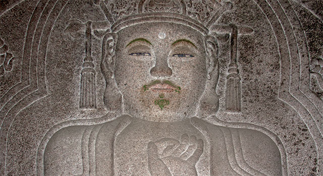Bas relief stone carving of a man with a serene expression, wearing a crown, and holding up his left hand with the tips of the thumb and index finger pressed together