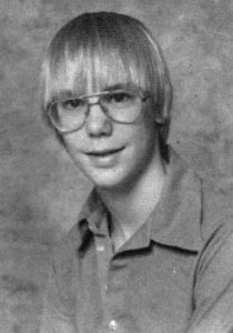 Black and white class photo of a 13 year old boy with glasses, a wide-collared shirt, and straight blond hair in long bangs